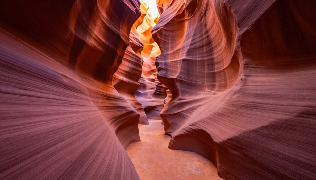 A stunning view of Antelope Canyon, Arizona, capturing the intricate, swirling sandstone formations and vibrant colors of the rock walls. In the foreground, smooth, sculpted rocks showcase shades of red, orange, and purple, illuminated by soft, golden sunlight filtering through the narrow canyon. The middle ground features a winding path that leads deeper into the canyon, accentuated by the play of light and shadows creating depth. The background reveals towering canyon walls that rise dramatically, with curves and arches forming a natural frame. The atmosphere is serene and ethereal, evoking a sense of wonder and exploration. This breathtaking landscape should be captured with a wide-angle lens to encompass the scale and beauty of the geological marvel, highlighting the textures and colors of the canyon's surface.
